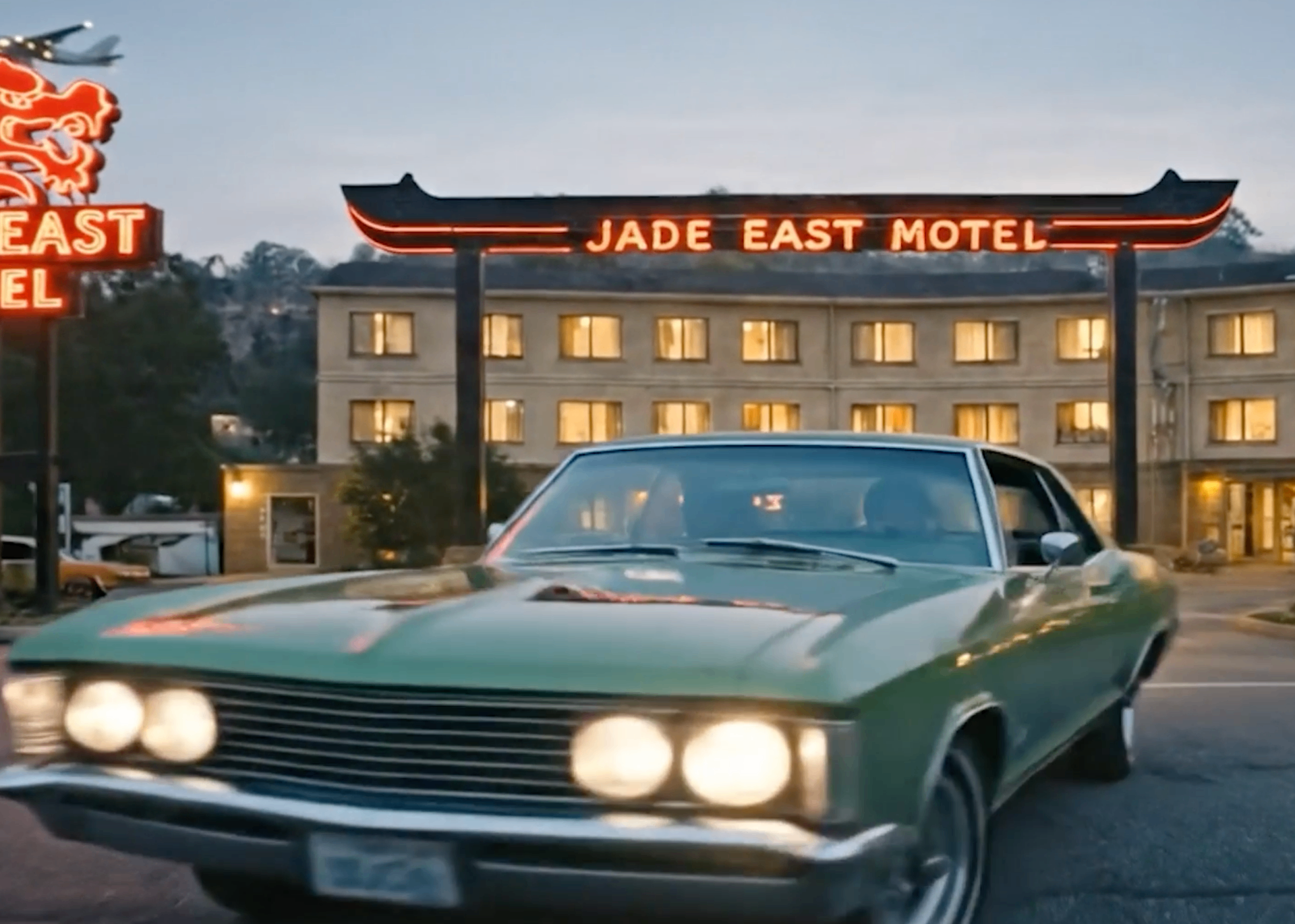 A green sedan idles in front of the neon-lit Jade East Motel at dusk